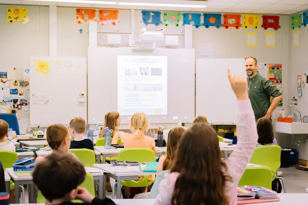 Mehrere Schüler im Unterricht in einem bunten Klassenraum. Vorne steht ein Lehrer und ein Mädchen meldet sich.
